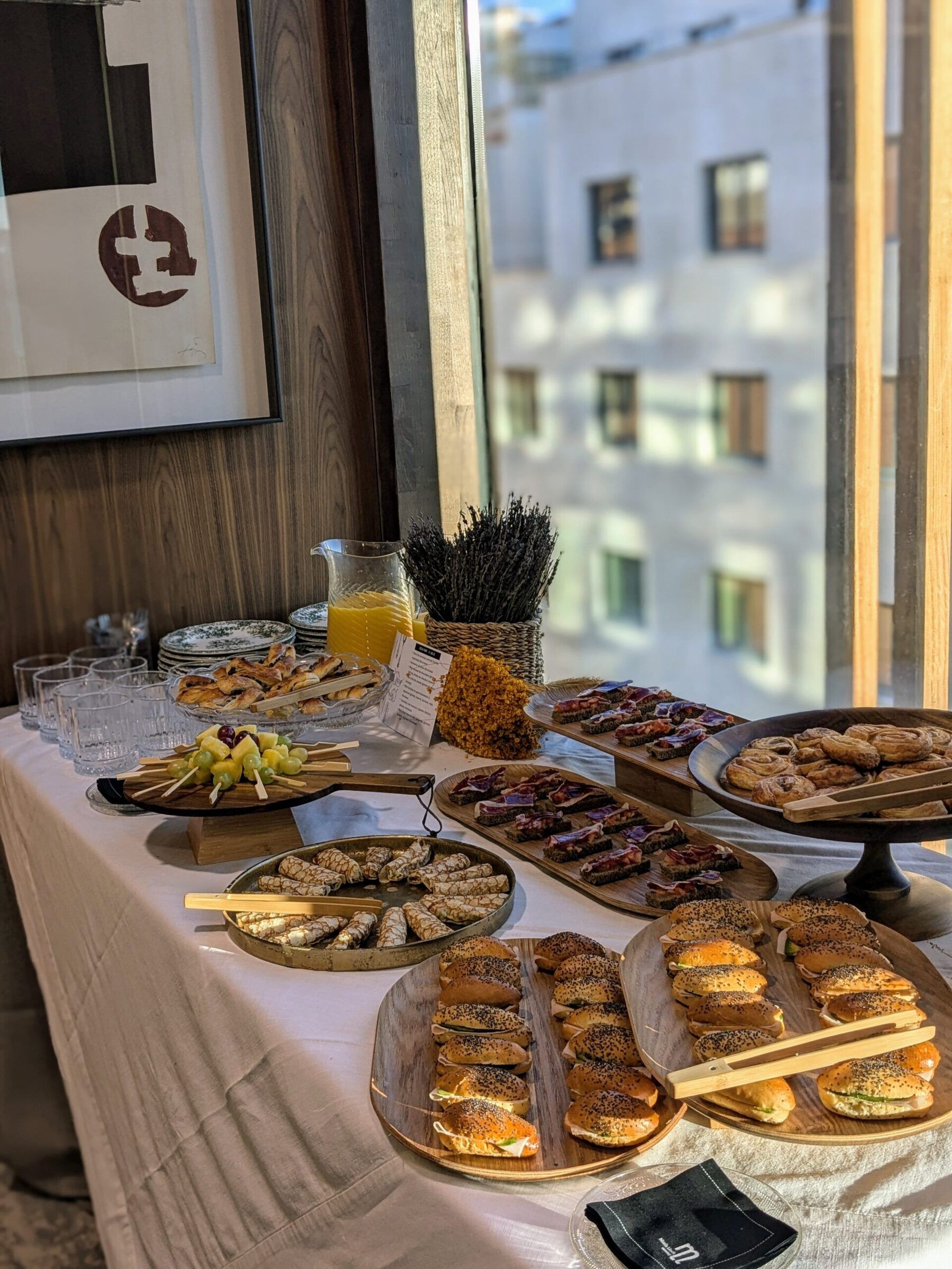 Office breakfast table by a large window with mini filled rolls, ham, pastries and orange juice, María Lluch catering in Madrid